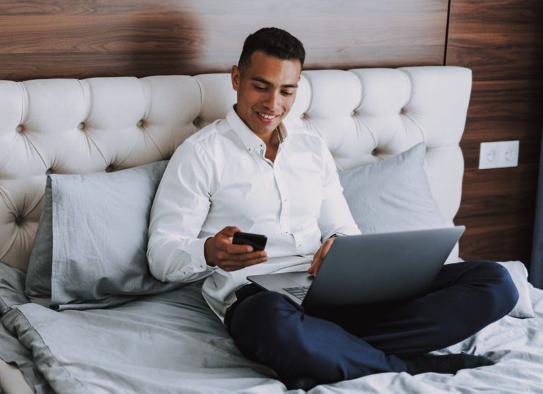 Man sitting on a bed with a laptop looking at his cell phone smiling.